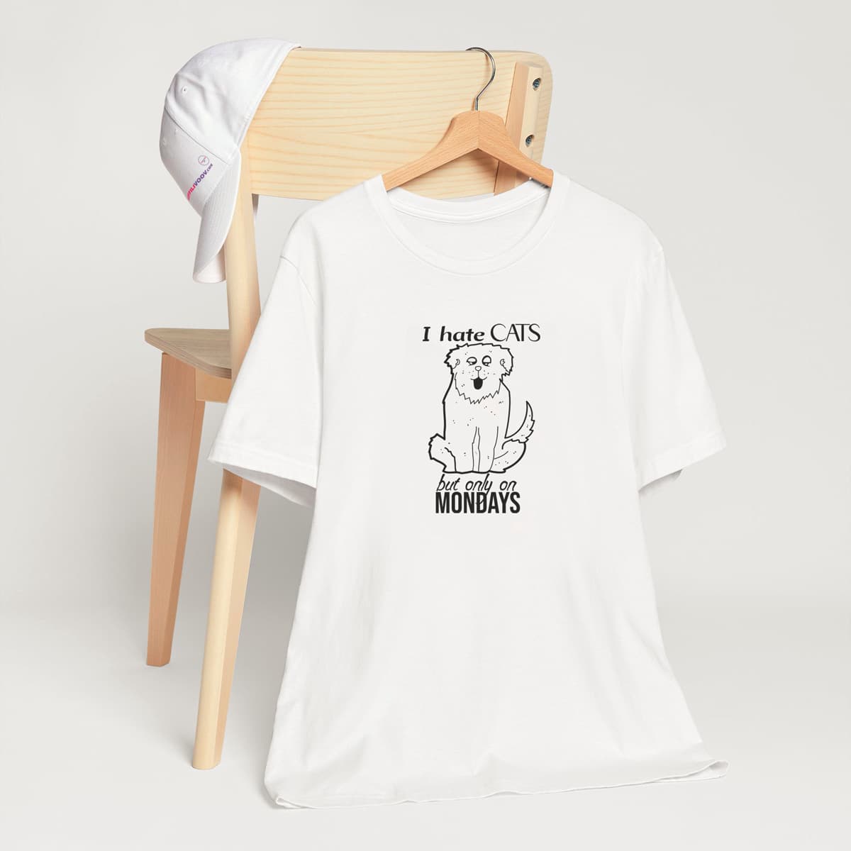 White crew neck short sleeved t-shirt, features a funny sitting dog, in bold black one-lined graphic with message ”I hate cats but only on Mondays”. T-shirt hanging on chair shown in close-up to highlight print and size