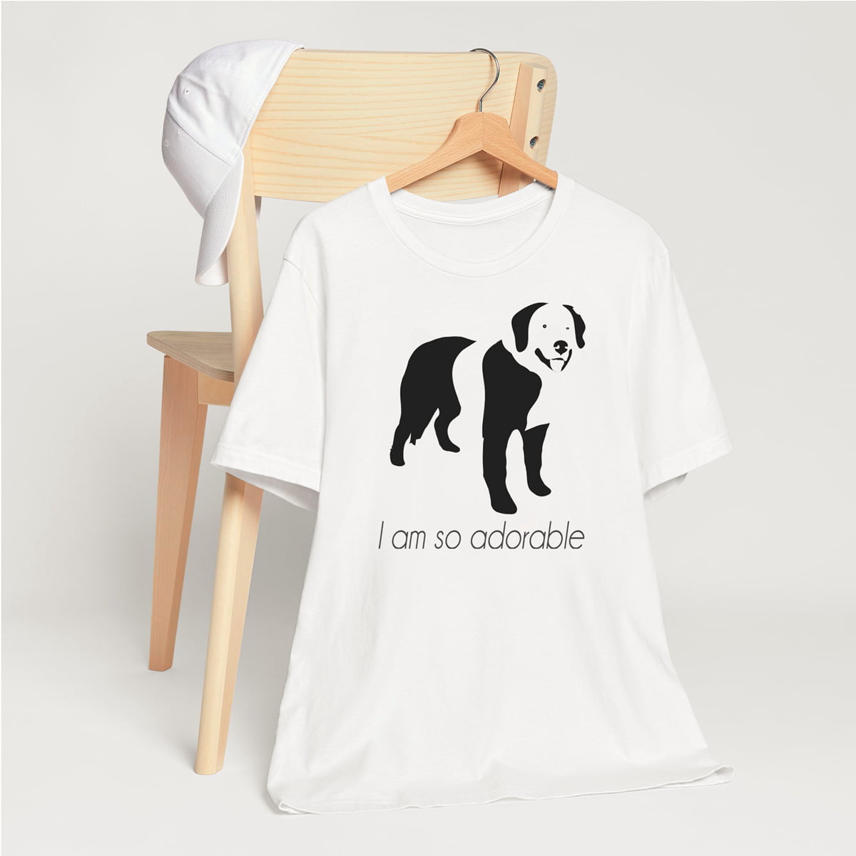 White crew neck short sleeved t-shirt, featuring a hand-drawn black and white one-line graphic print silhouette of an adorable Bernese Mountain with its tongue out. The text "I am so adorable" underneath the dog .T-shirt hanging on chair shown in close-up to highlight print and size