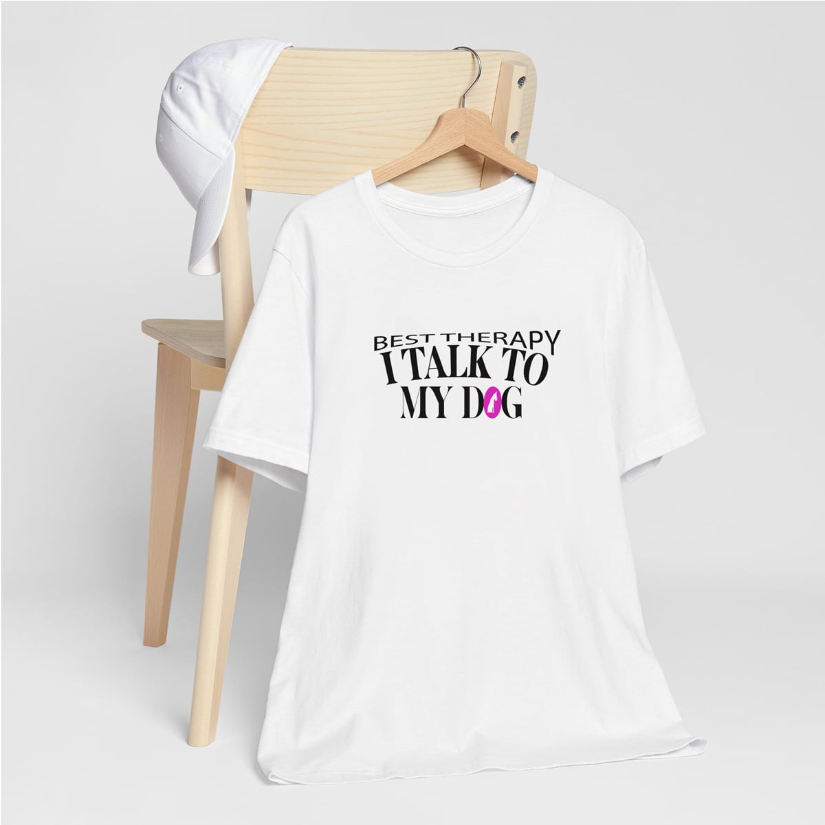 White crew neck short sleeved t-shirt, feature a graphic ”best therapy i talk to my dog” quote a cross chest. T-shirt hanging on chair shown in close-up to highlight print and size.