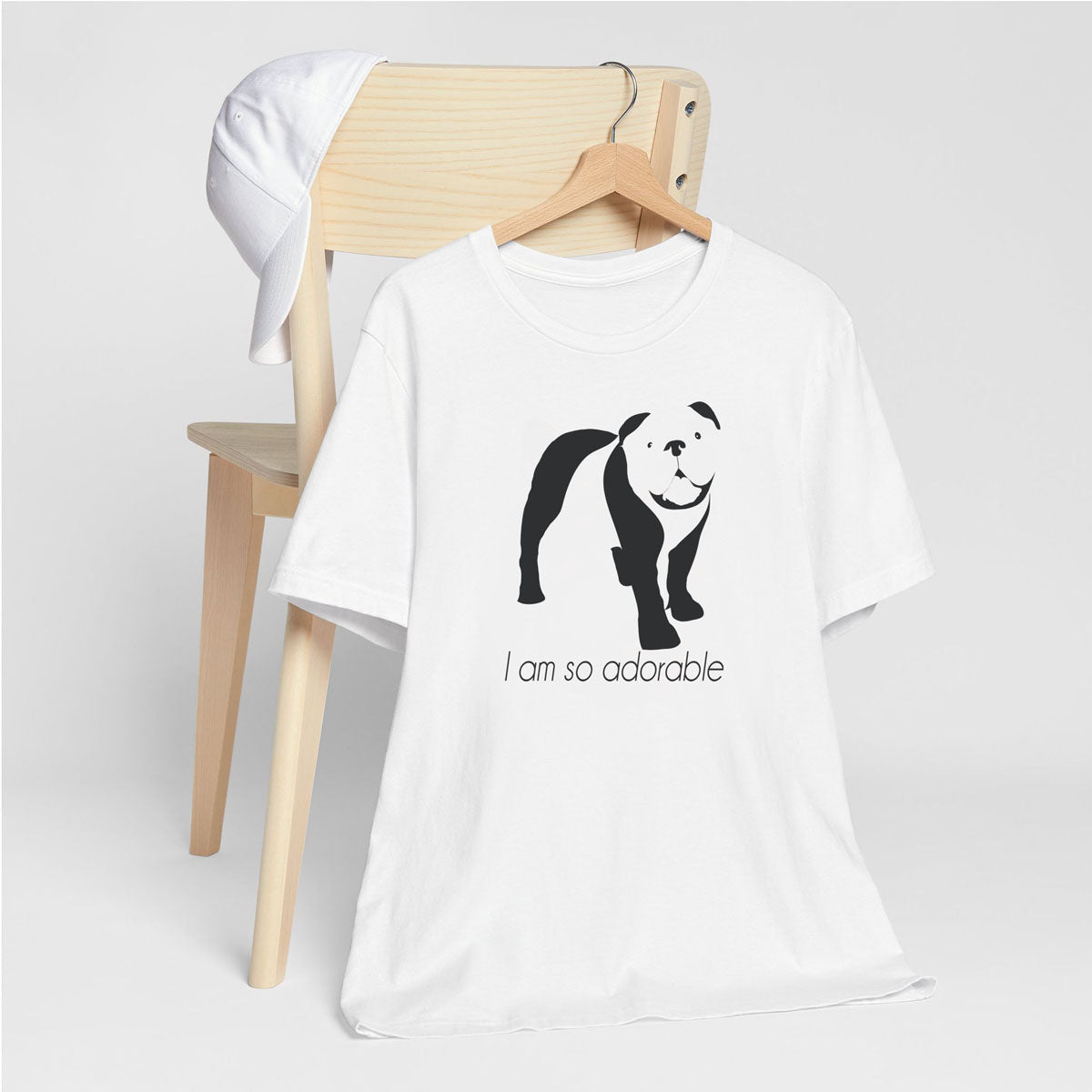 White crew neck short sleeved t-shirt, features a charming bulldog silhouette with playful quote; I am so adorable” across chest. T-shirt hanging on chair shown in close-up to highlight print and size.