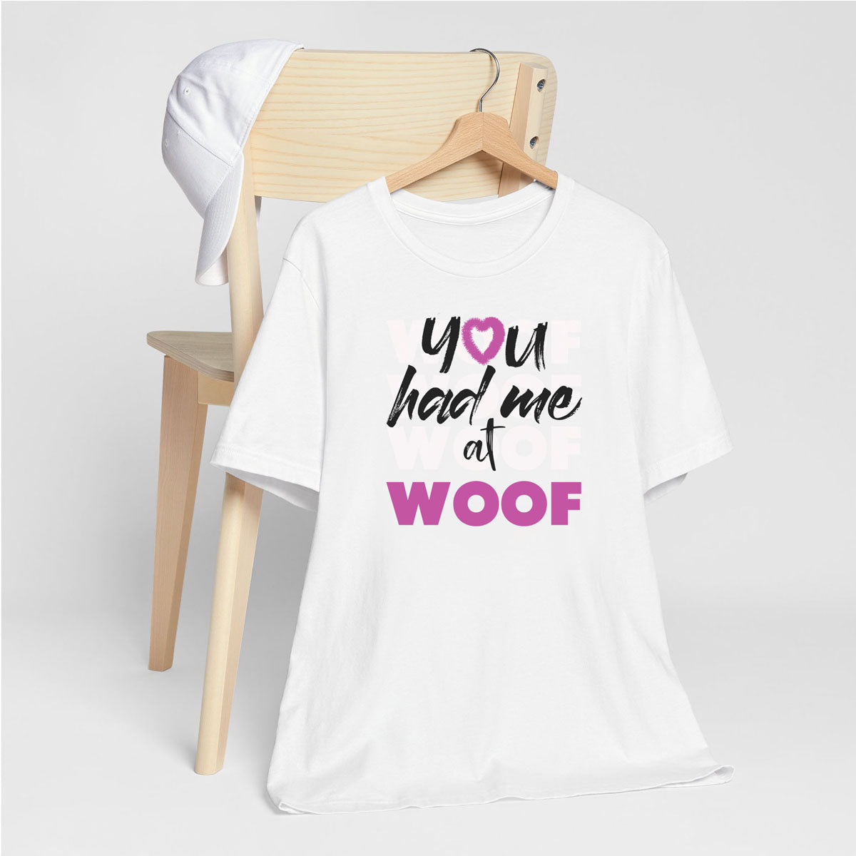 White crew neck short sleeved t-shirt, feature a graphic classic quote ”you had me at woof” across chest. T-shirt hanging on chair shown in close-up to highlight print and size.