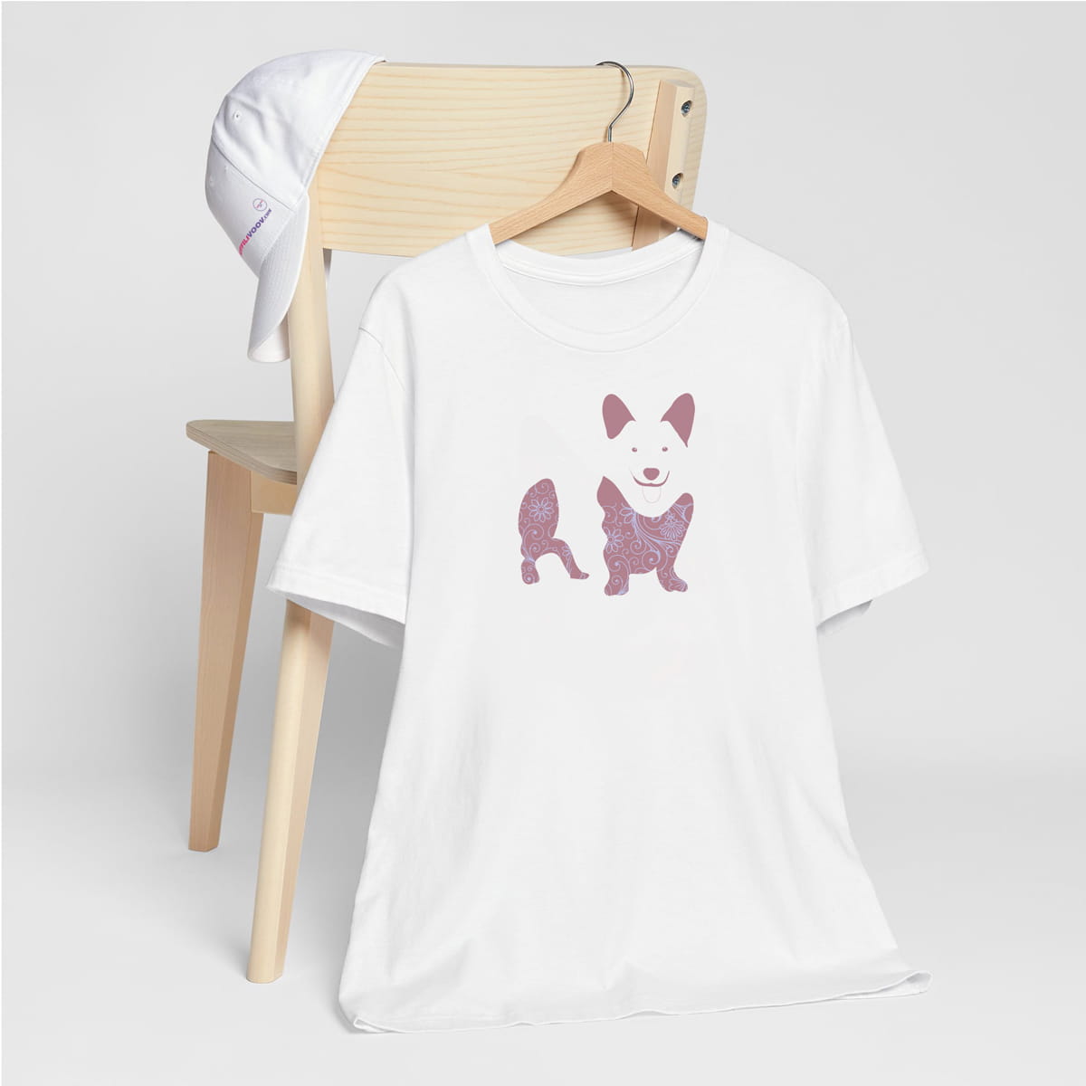 White crew neck short sleeved t-shirt, features a charming Corgi silhouette with floral, and patterned design. hanger on chair. T-shirt hanging on chair shown in close-up to highlight print and size