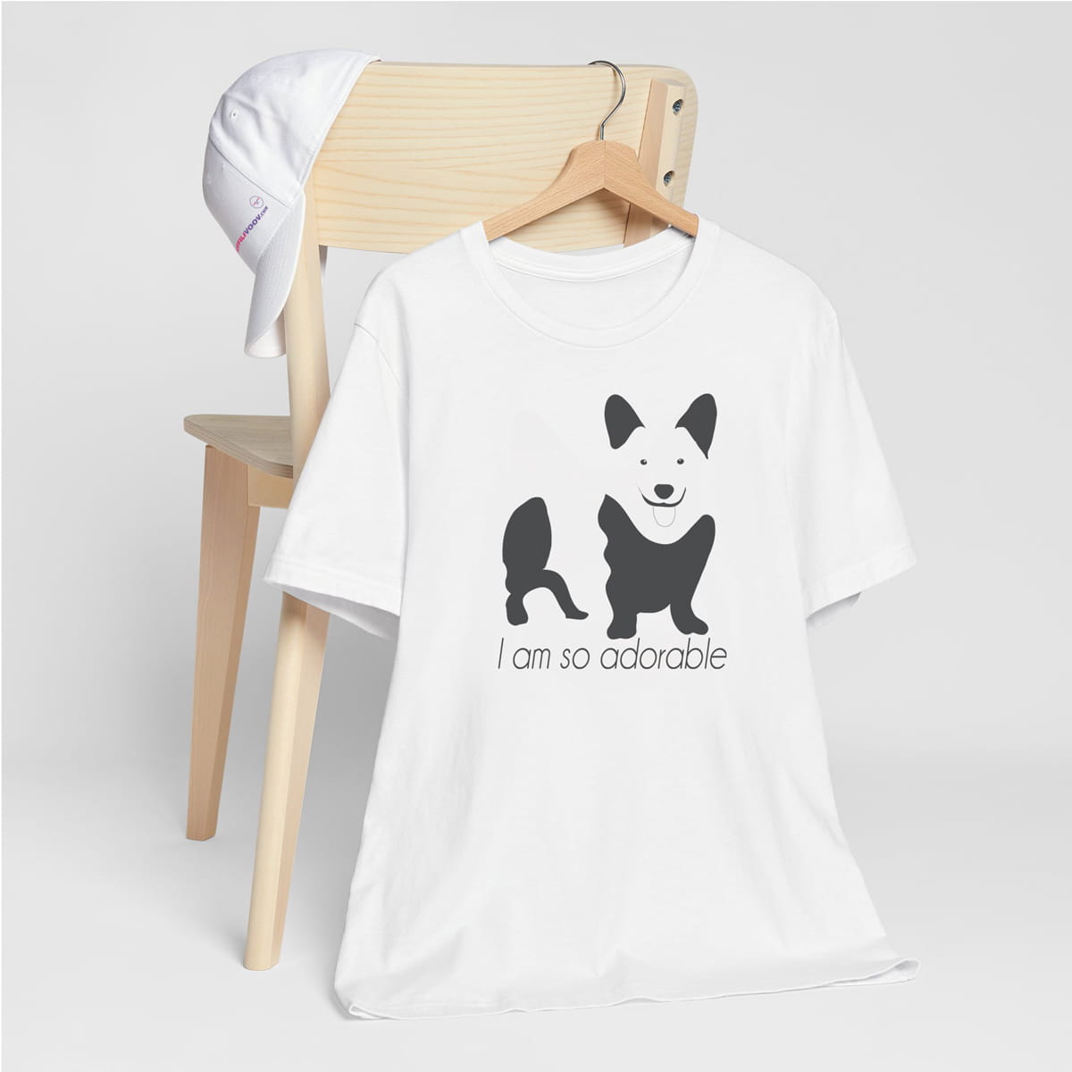 White crew neck short sleeved t-shirt, feature a graphic black/white hand-drawn corgi standing dog with quote " i am so adorable". T-shirt hanging on chair shown in close-up to highlight print and size.