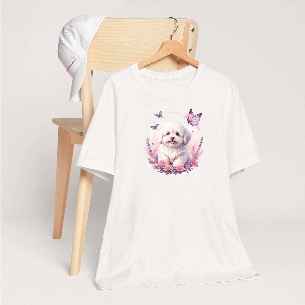 White crew neck short sleeve t-shirt with a colorful hand-drawn Coton de tulear puppy graphic sitting in a floral garden. t-shirt hanging on chair, close up showing bold dog art, cute pet-themed fashion