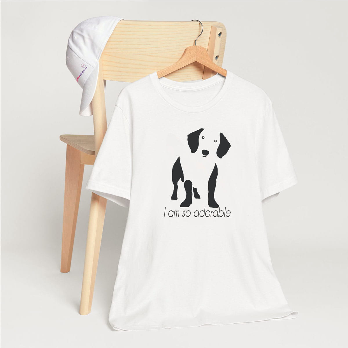 White crew neck short sleeved t-shirt, feature a feature a standing dachshund in black/white silhouette with quote "I am so adorable". T-shirt hanging on chair shown in close-up to highlight print and size