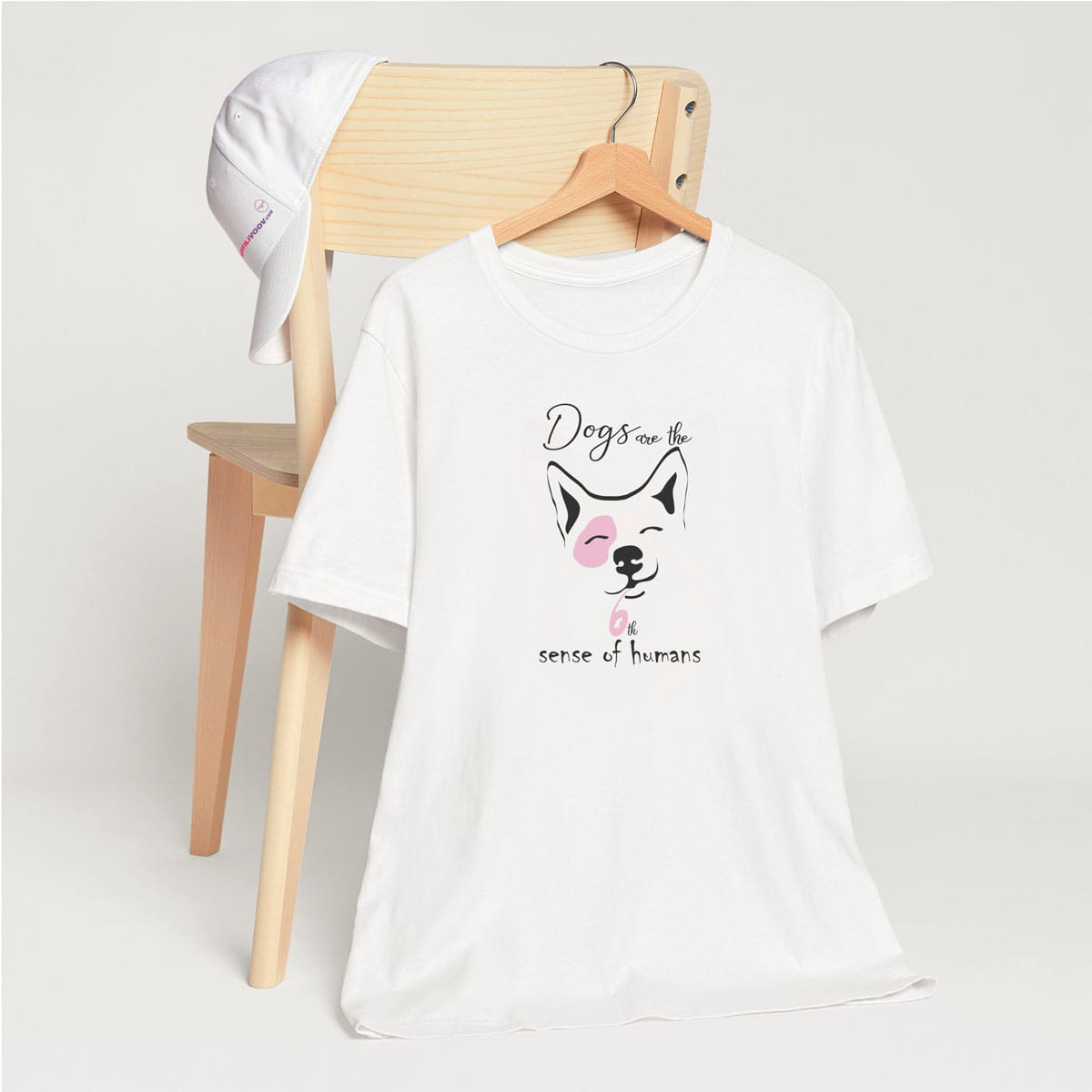 White crew neck short sleeved t-shirt, feature an adorable corgi face squint-eyed pooch backed by the humorous text "dogs are the 6th sense of humans. T-shirt hanging on chair shown in close-up to highlight print and size