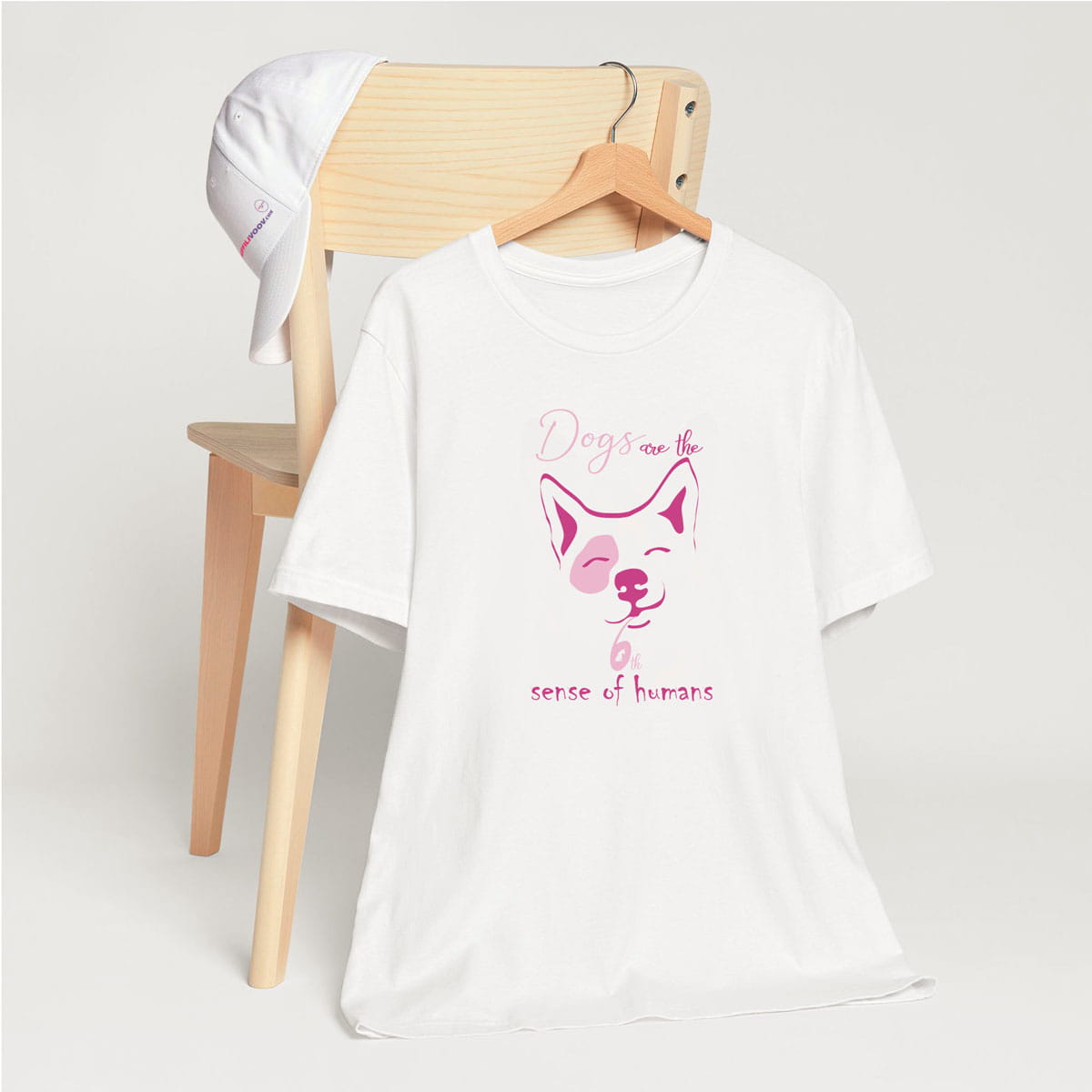 White crew neck short sleeved t-shirt, feature a cute happy corgi dog face, with a fun statement, quote with a twist ”Dogs are the 6th sense of humans”. T-shirt hanging on chair shown in close-up to highlight print and size.