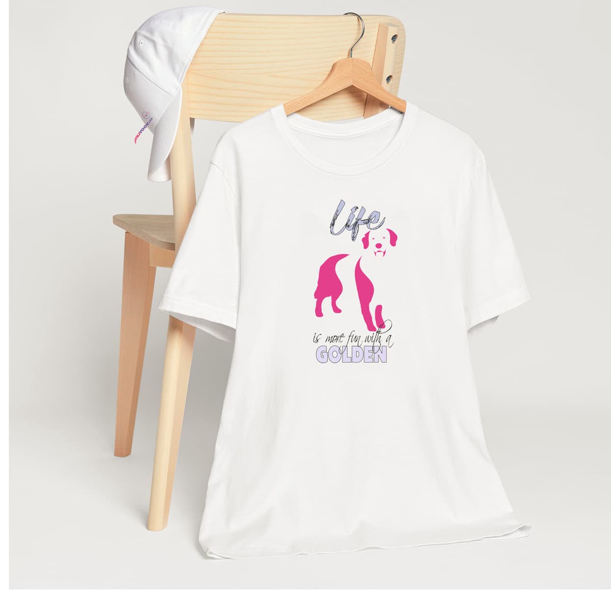 White crew neck short sleeved t-shirt. Featuring an adorable Golden Retriever silhouette in cute pink, the tee also boasts a bold "Life" message and empowering "is more fun with a Golden" phrase. T-shirt hanging on chair shown in close-up to highlight print and size.
