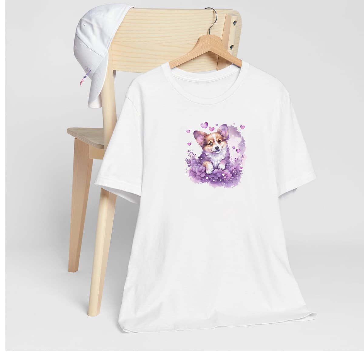 White crew neck short sleeved t-shirt, features a cute Corgi puppy dog nestle in hearts and floral garden, in a burst vibrant warm colors. T-shirt hanging on chair shown in close-up to highlight print and size