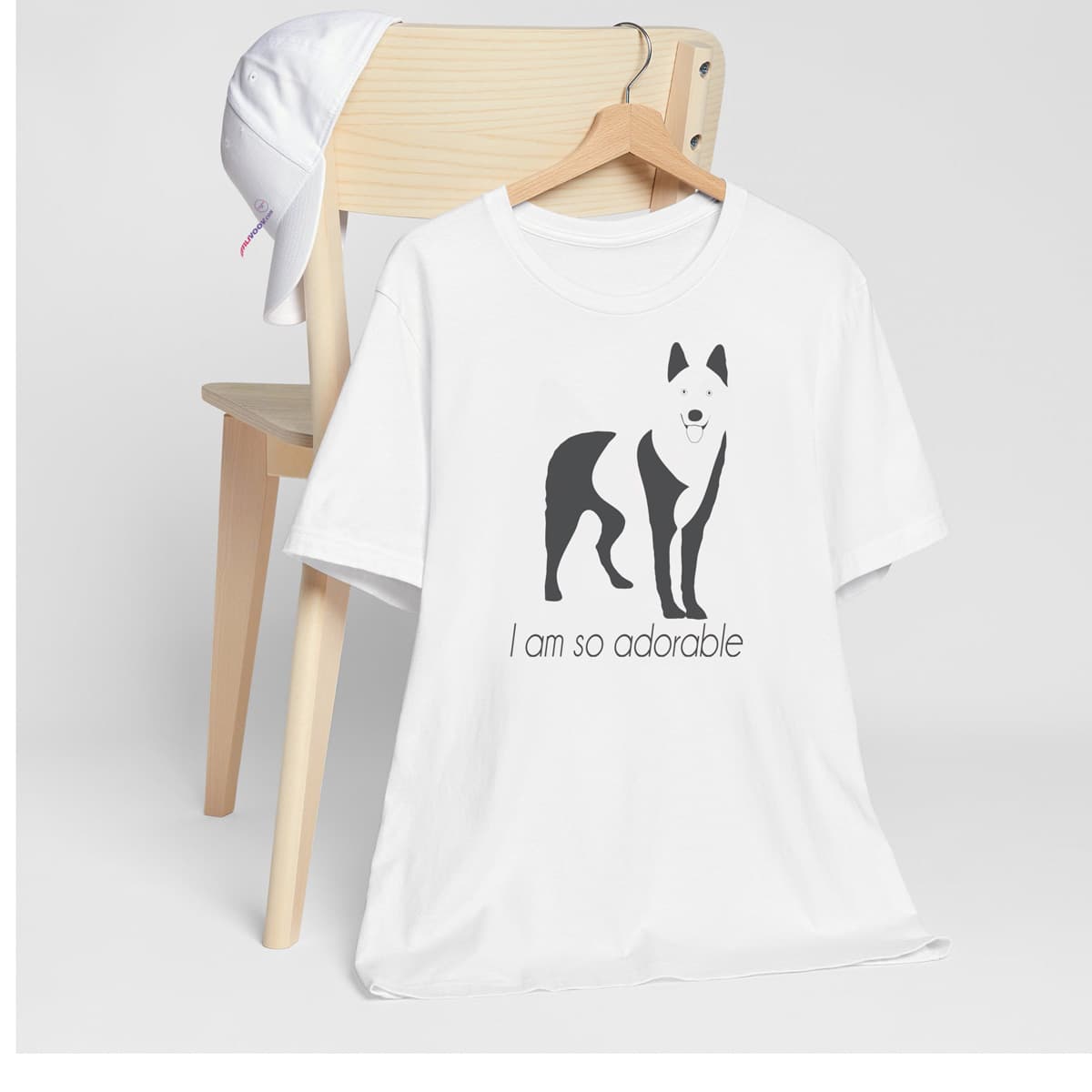 White crew neck short sleeved t-shirt, featuring a one-line graphic print of a hand-drawn cute Siberian Husky with its tongue out, and the playful text "I am so adorable”. T-shirt hanging on chair shown in close-up to highlight print and size