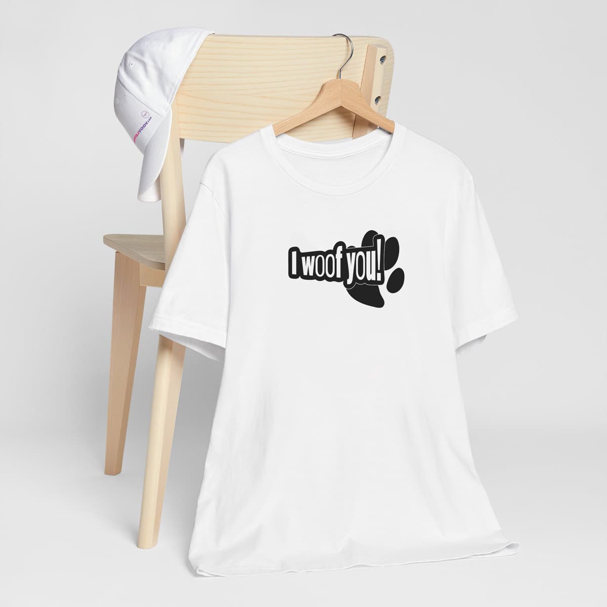 White crew neck short sleeved t-shirt, feature a funny message "I woof you" designed in a cartoonist way, underlined by a dog paw. T-shirt hanging on chair shown in close-up to highlight print and size.