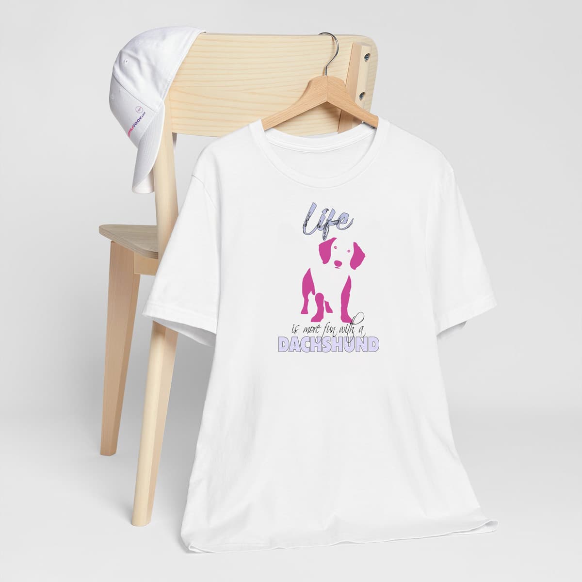 White crew neck short sleeved t-shirt. Featuring a silhouette Dachshund in vibrant pink, and quote with humor "life is more fun with a Dachshund"T-shirt hanging on chair shown in close-up to highlight print and size.