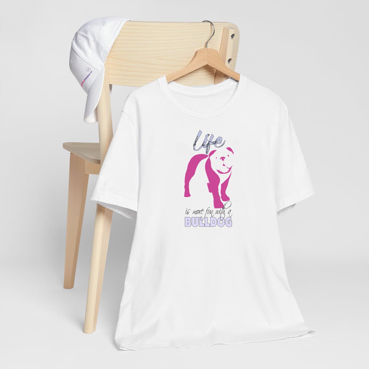 White crew neck short sleeved t-shirt. Featuring an adorable Bulldog silhouette in cute pink, the tee also boasts a bold "Life" message and empowering "is more fun with a Golden" phrase. T-shirt hanging on chair shown in close-up to highlight print and size
