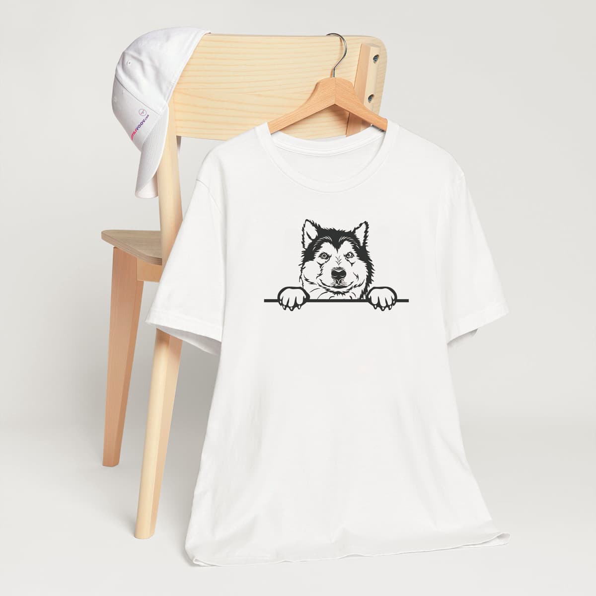 White crew neck short sleeved t-shirt. Showcase an adorable black/white hand-drawn graphic Malamute dog face resting on a table. T-shirt hanging on chair shown in close-up to highlight print and size.