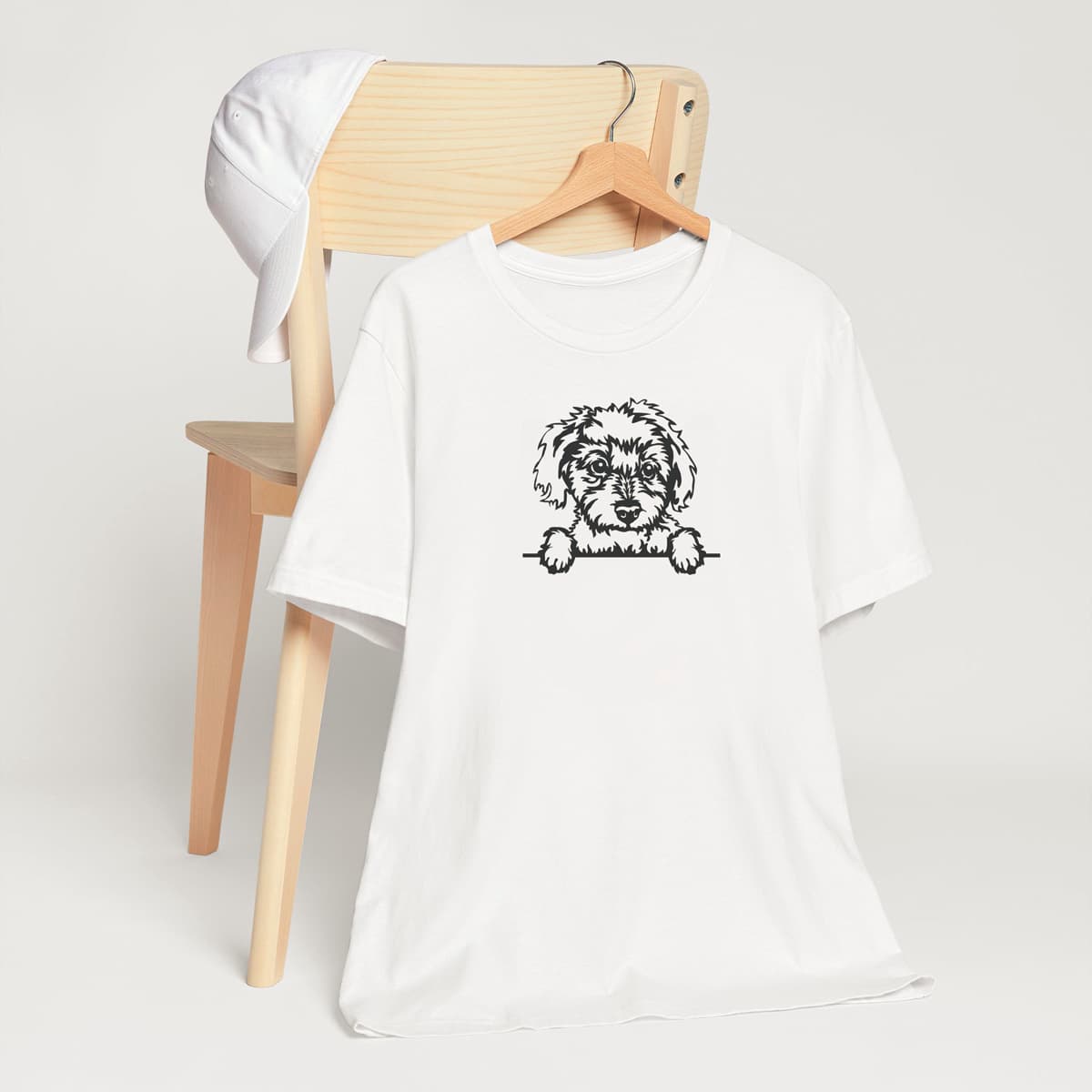 White crew neck short sleeved t-shirt. Feature a charming black and white one-line graphic of a Maltese dog face with paws on the table, and a playful "no Peeking" scheme.T-shirt hanging on chair shown in close-up to highlight print and size.