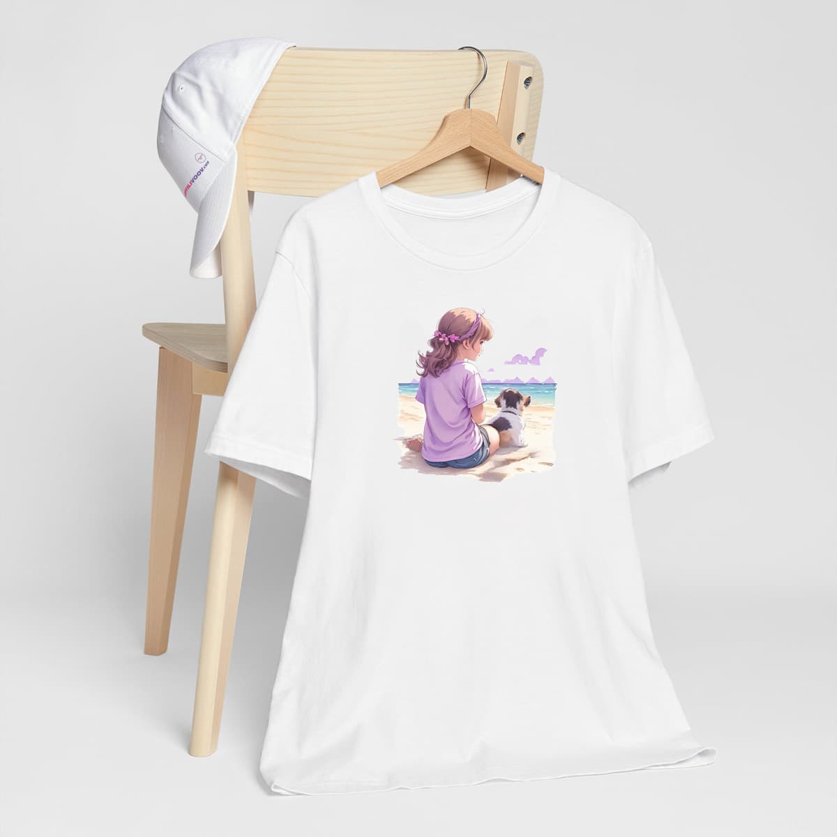 White crew neck short sleeved t-shirt ,feature a watercolor hand-drawn illustration of a girl and her adorable puppy, frolicking on a beach vista against an ocean backdrop. T-shirt hanging on chair shown in close-up to highlight print and size