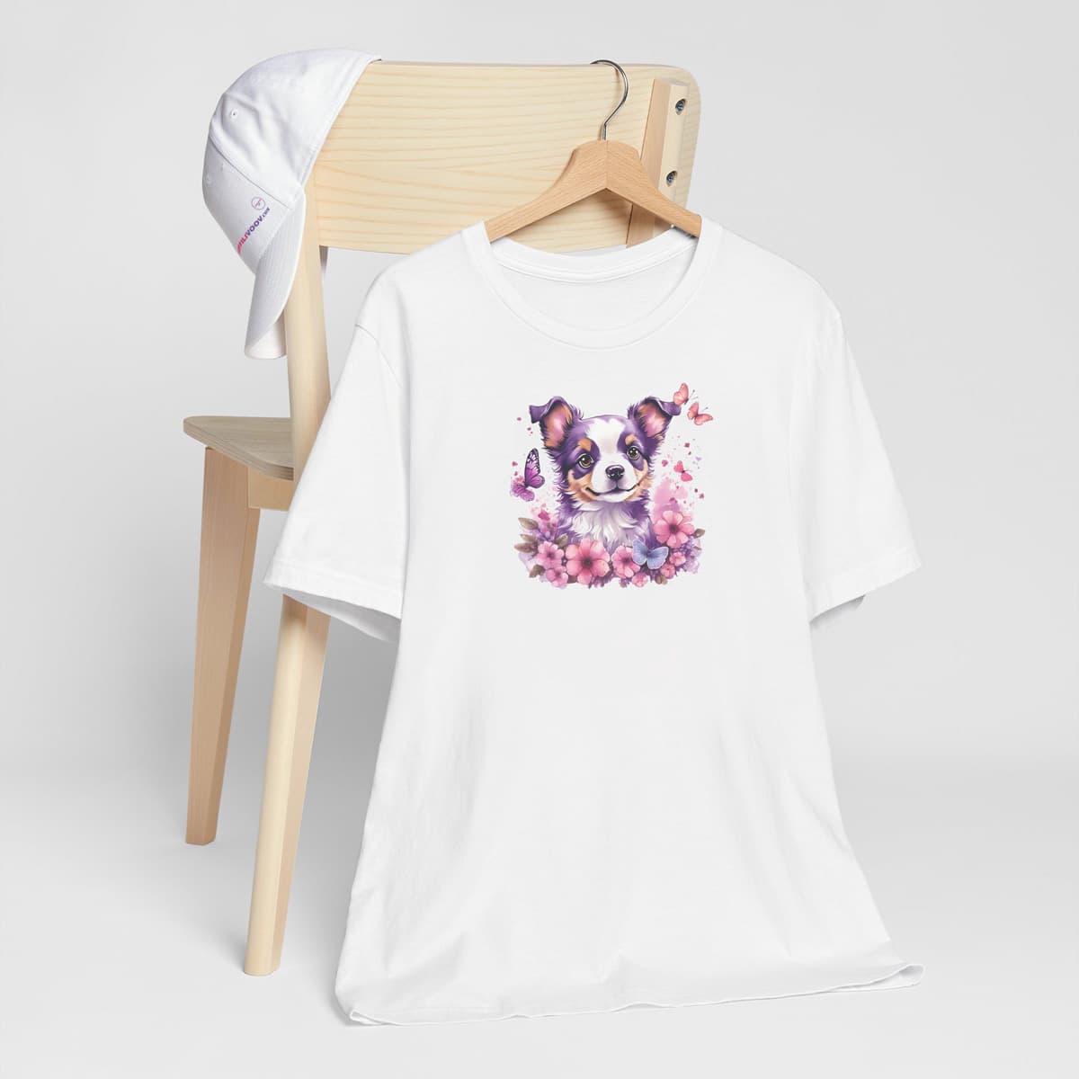 White crew neck short sleeved t-shirt, showcases a puppy face, nestled amidst a lush floral arrangement within a wicker basket. T-shirt hanging on chair shown in close-up to highlight print and size