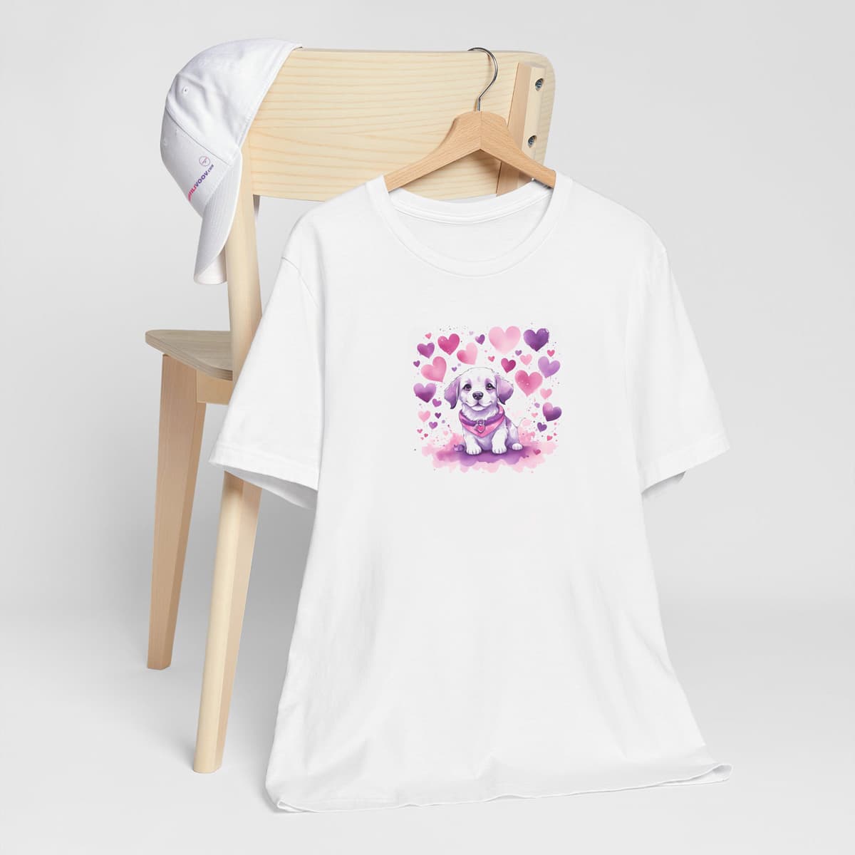 White crew neck short sleeved t-shirt. This cute shirt, radiate off colorful hearts, and a standing happy puppy in warmth toned palette. T-shirt hanging on chair shown in close-up to highlight print and size