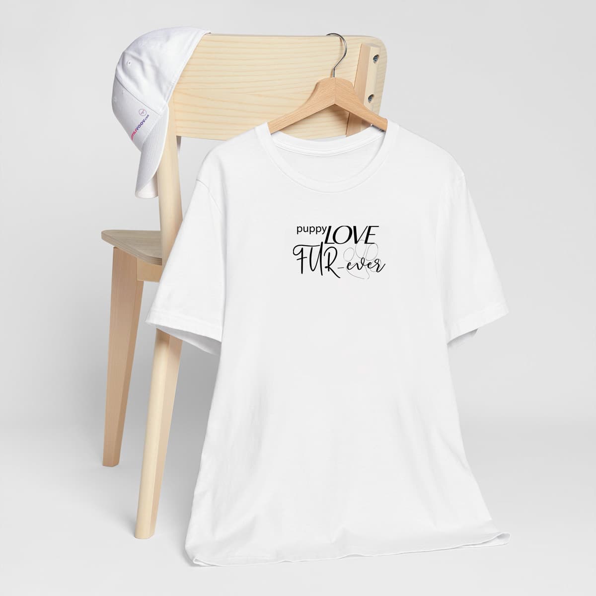 White crew neck short sleeved t-shirt, feature a message "puppy love fur-ever" underlined with a dog paw. T-shirt hanging on chair shown in close-up to highlight print and size.