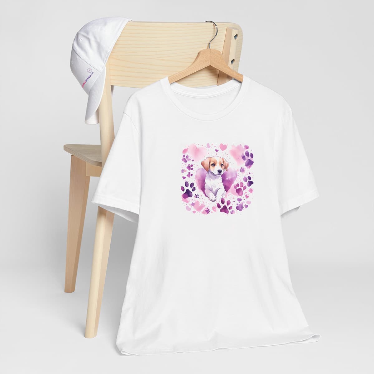White crew neck short sleeved t-shirt, Feature a vibrant colorful design of a lively, adorable puppy surrounded by a whirlwind of paws and hearts. T-shirt hanging on chair shown in close-up to highlight print and size