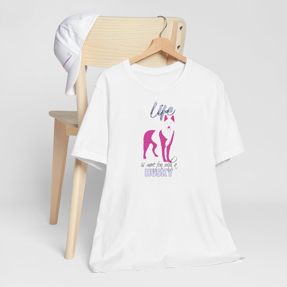 White crew neck short sleeved t-shirt. Featuring an adorable Siberian Husky silhouette in cute pink, the tee also boasts a bold "Life" message and empowering "is more fun with a Husky" phrase.T-shirt hanging on chair shown in close-up to highlight print and size