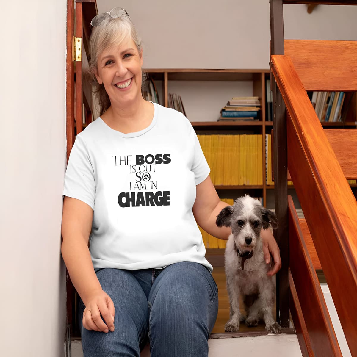 Women sitting at a indoor staircase, with a small dog. She is wearing a white crew neck short sleeved t-shirt, features a playful message "the boss is out so i am in charge" in bold black graphic style, underlined by a dog paw