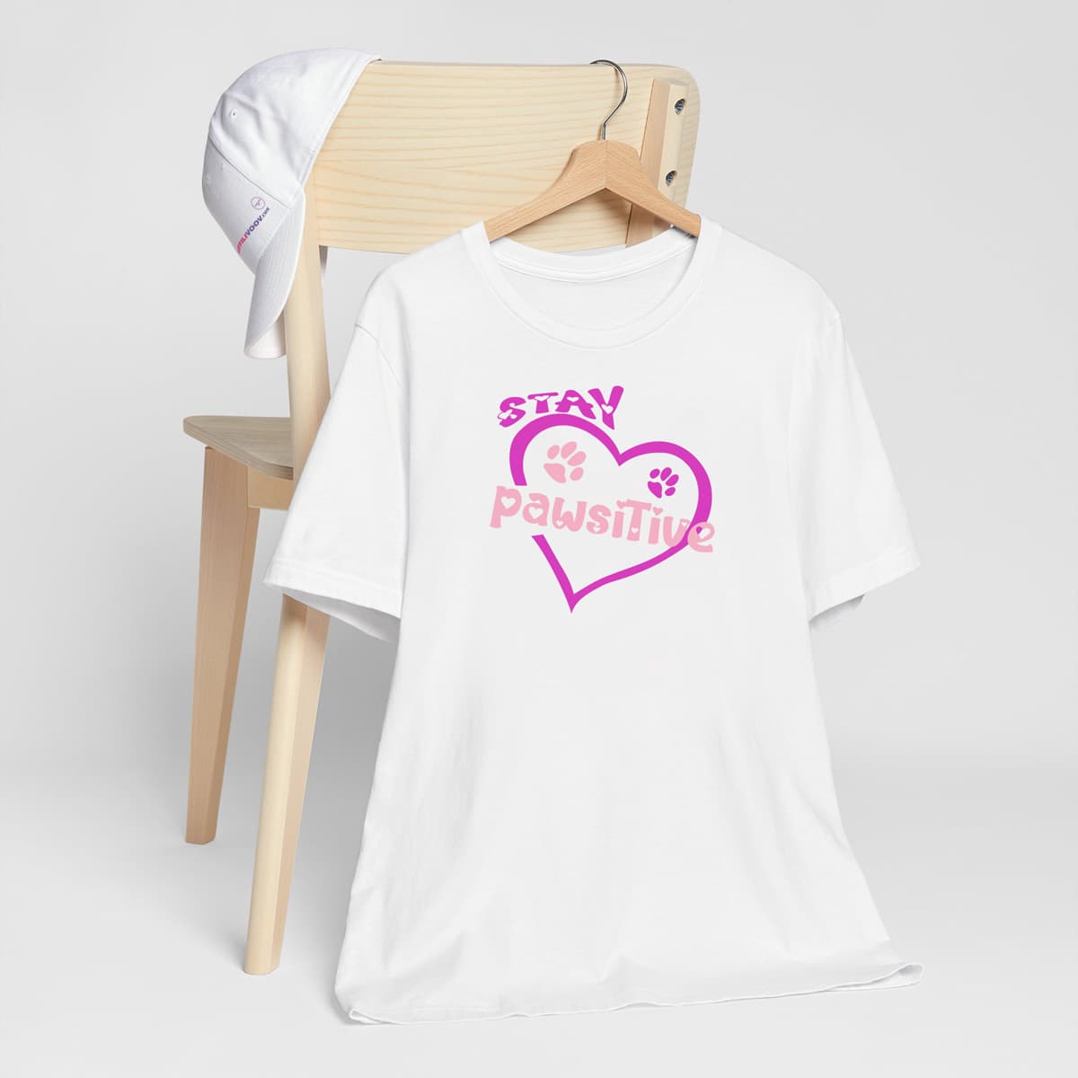 White crew neck short sleeved t-shirt, featuring pink and purple paws nestle in a pink heart and a fun message ”stay paw-sitive” with doggo twist in bold vibrant colorful graphic style.T-shirt hanging on chair shown in close-up to highlight print and size