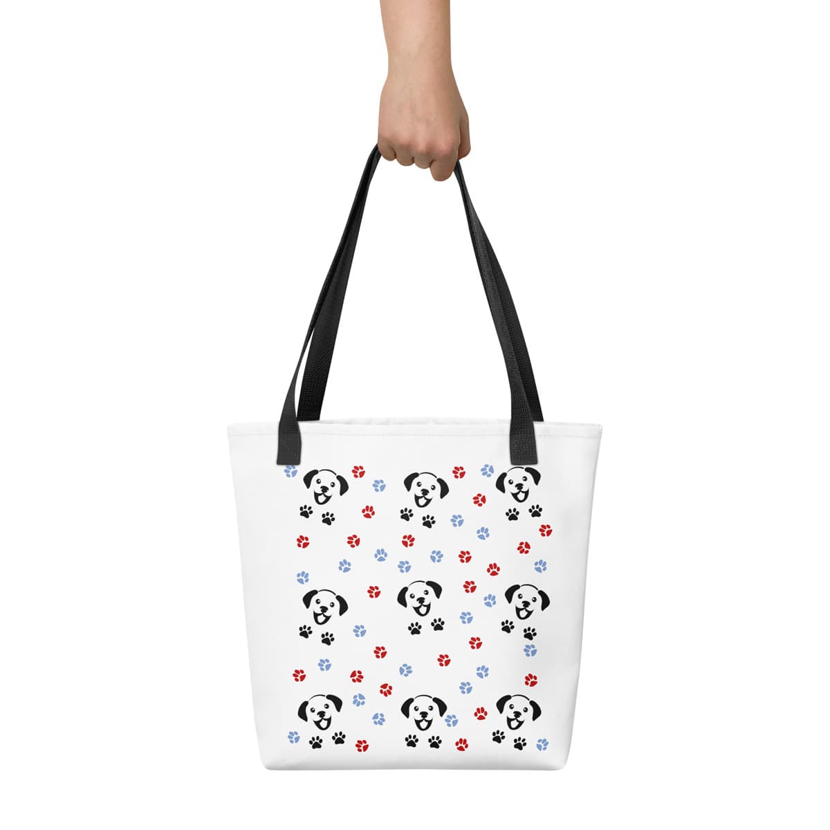 Hand holding a white spun canvas fabric tote bag, with black handle stripped, on the tote is a cute graphic cartoonist happy puppy face, surrounded by colorful paws. Hand model showing front of tote bag.