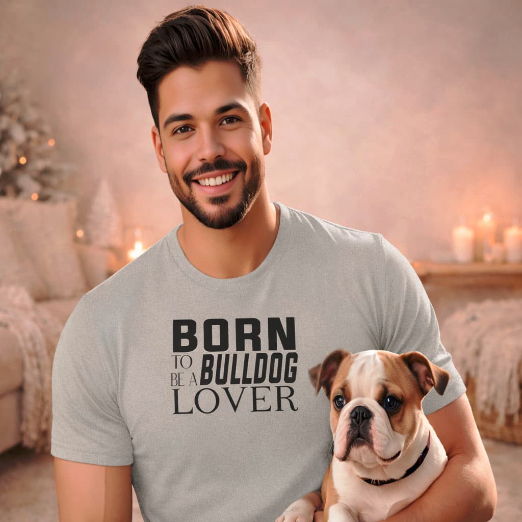 Smiling man holding a bulldog puppy indoors, wearing a gray feature Born to be a bulldog lover text graphic short sleeve t-shirt in a warm winter studio interior with soft daylight and neutral tones.