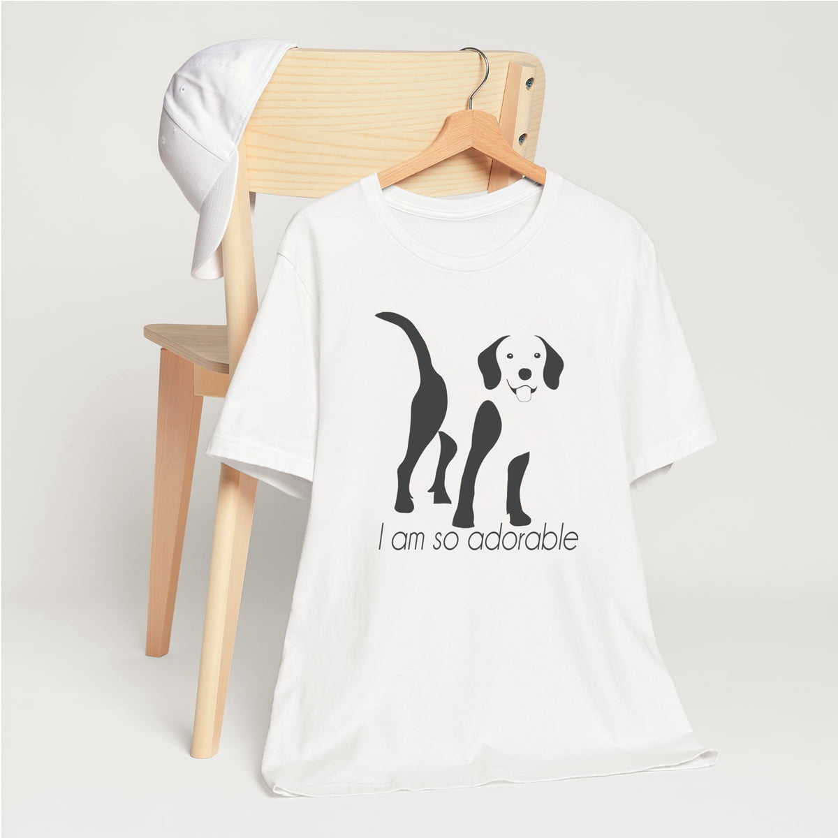 White crew neck short sleeved t-shirt, featuring an adorable Beagle hand drawn silhouette graphic print with the text "I am so adorable." underneath the dog. T-shirt hanging on chair shown in close-up to highlight print and size