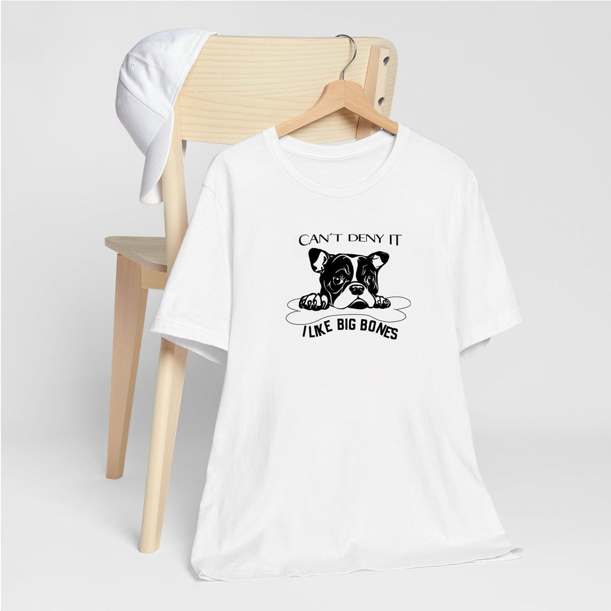 White crew neck short sleeved t-shirt, feature a graphic black/white hand-drawn cute bulldog face protecting its dog bone. T-shirt hanging on chair shown in close-up to highlight print and size