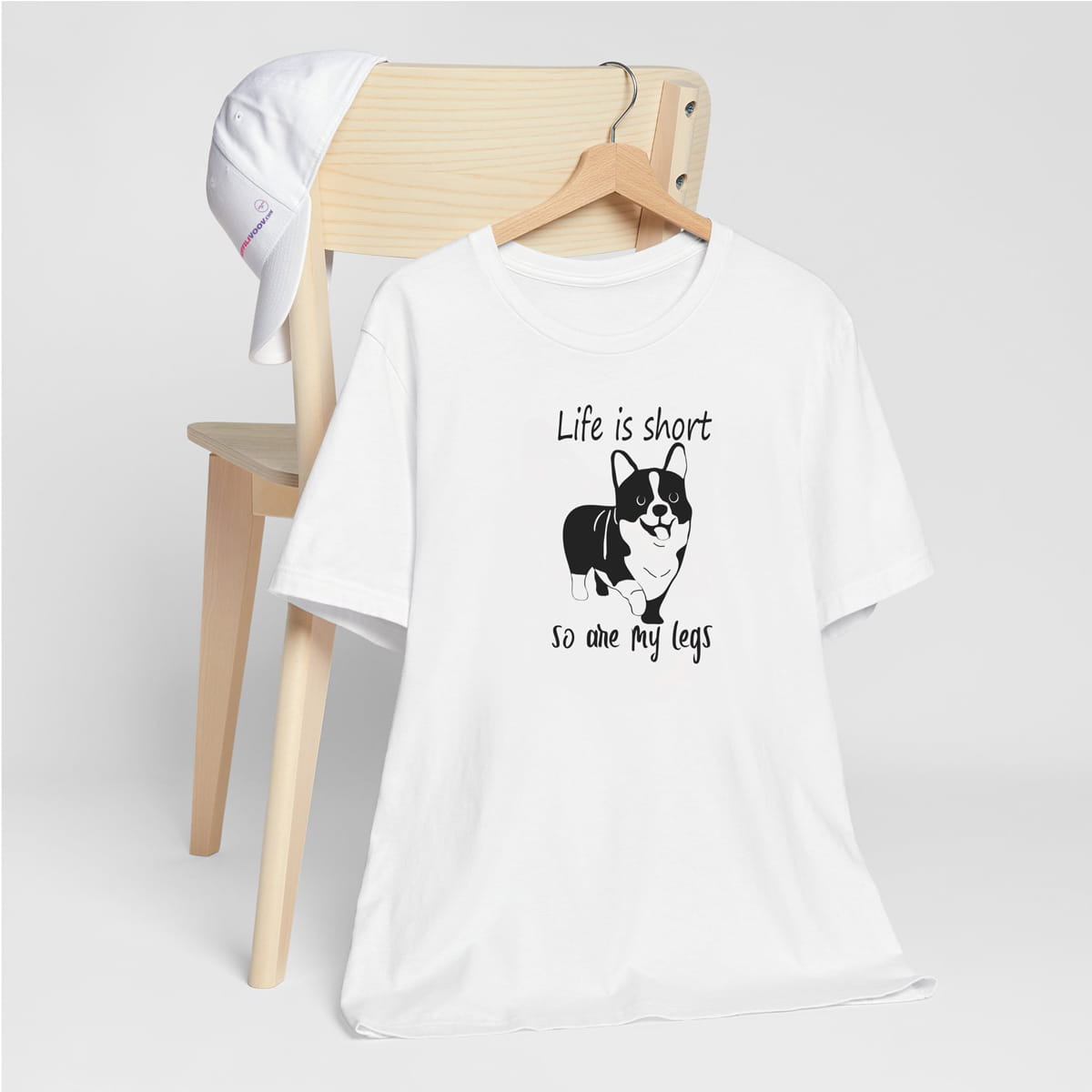 White crew neck short sleeved t-shirt, feature a graphic black/white hand drawn Corgi and text ”life is short so are my legs” quote across chest. T-shirt hanging on chair shown in close-up to highlight print and size
