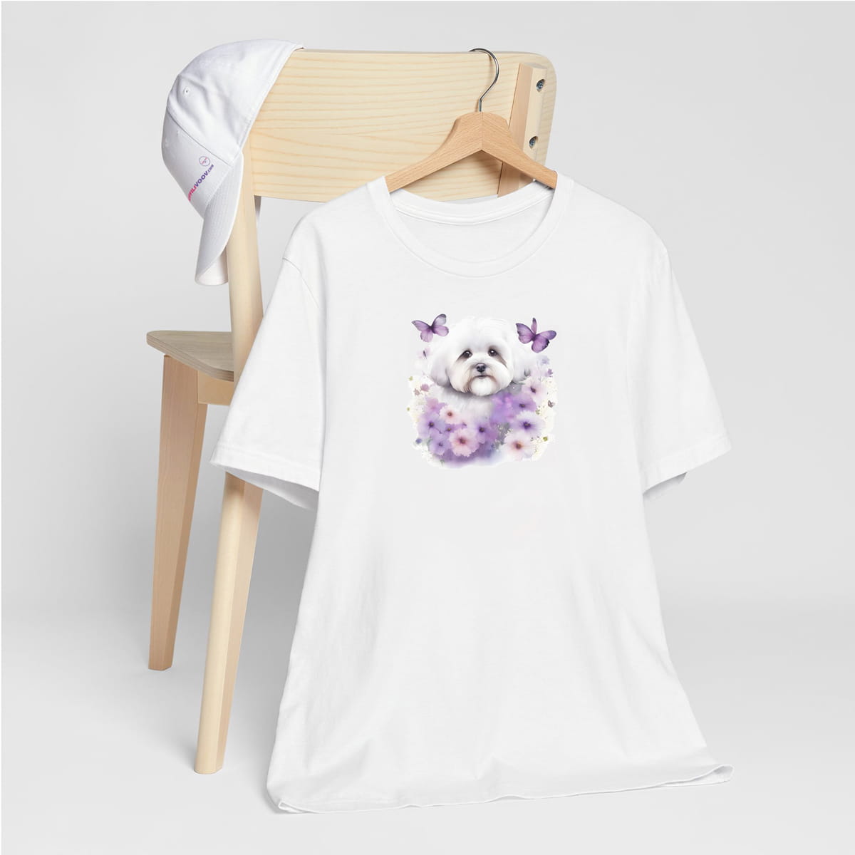 White crew neck short sleeve t-shirt with a colorful hand-drawn Coton de tulear puppy graphic sitting in a floral garden. shirt, hanging on chair close up showing bold dog art, cute pet-themed fashion