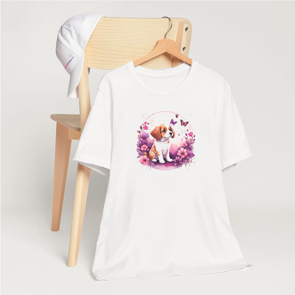 White crew neck short sleeved t-shirt, feature a playful pup amidst a whimsical floral fiesta. T-shirt hanging on chair shown in close-up to highlight print and size