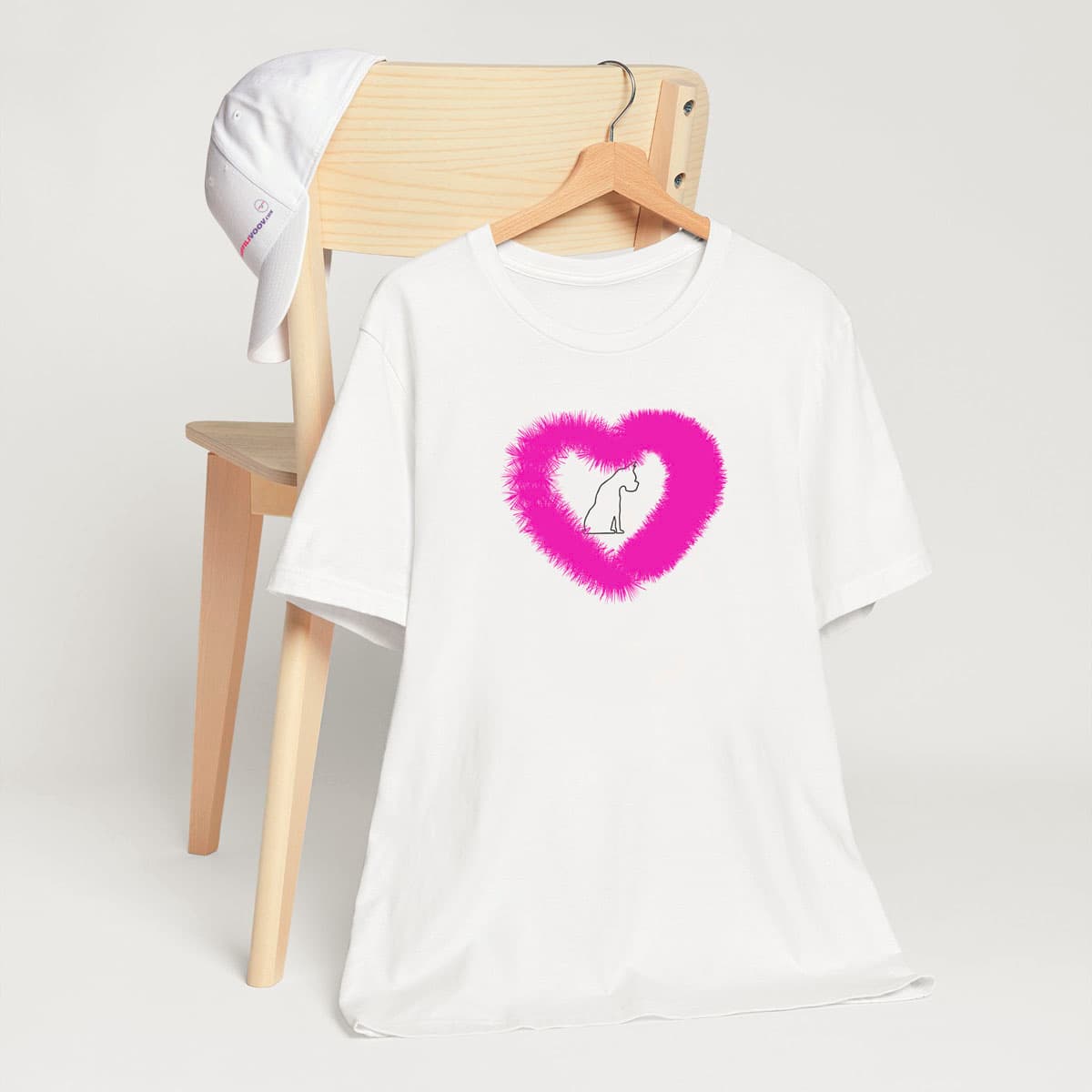 White crew neck short sleeved t-shirt, feature a sitting black one-lined hand drawn dog, in a fluffy pink heart.T-shirt hanging on chair shown in close-up to highlight print and size.
