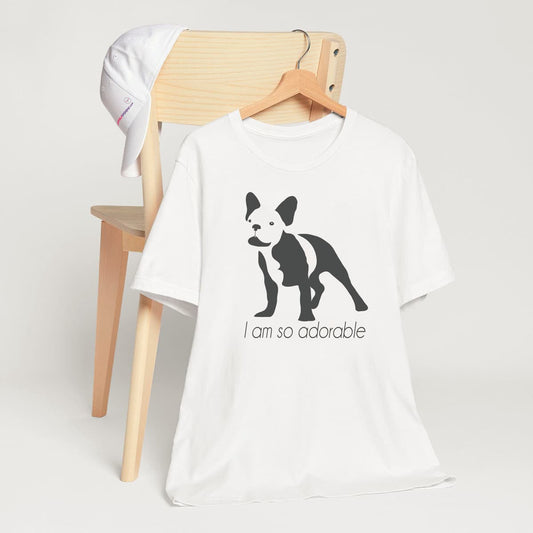 White crew neck short sleeved t-shirt, featuring a one-line graphic print of a hand-drawn French Bulldog with its tongue out, and the playful text "I am so adorable”. T-shirt hanging on chair shown in close-up to highlight print and size.