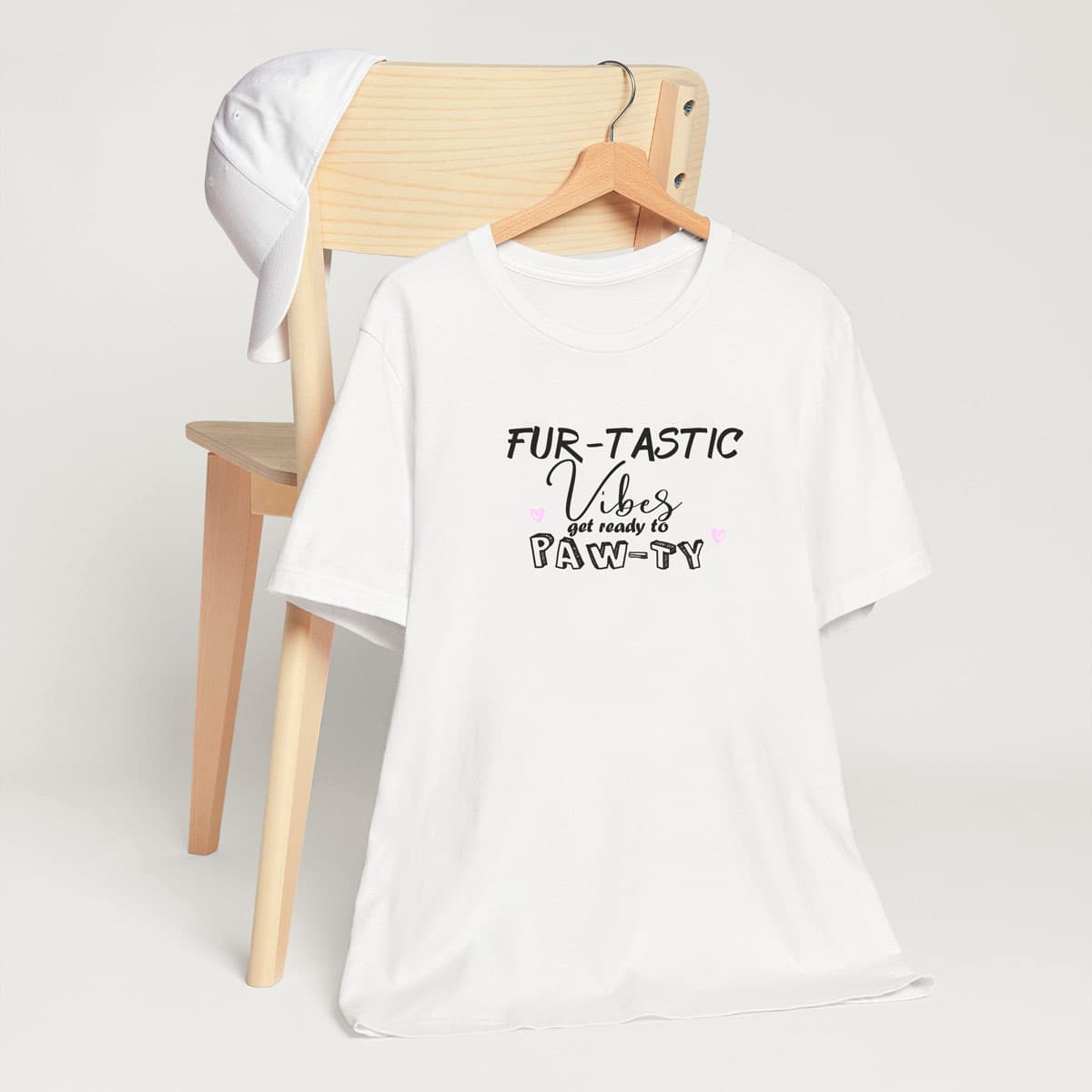 White crew neck short sleeved t-shirt, features a fun message "fur-tastic vibes get ready to paw-ty black graphic with cute pink hearts. T-shirt hanging on chair shown in close-up to highlight print and size