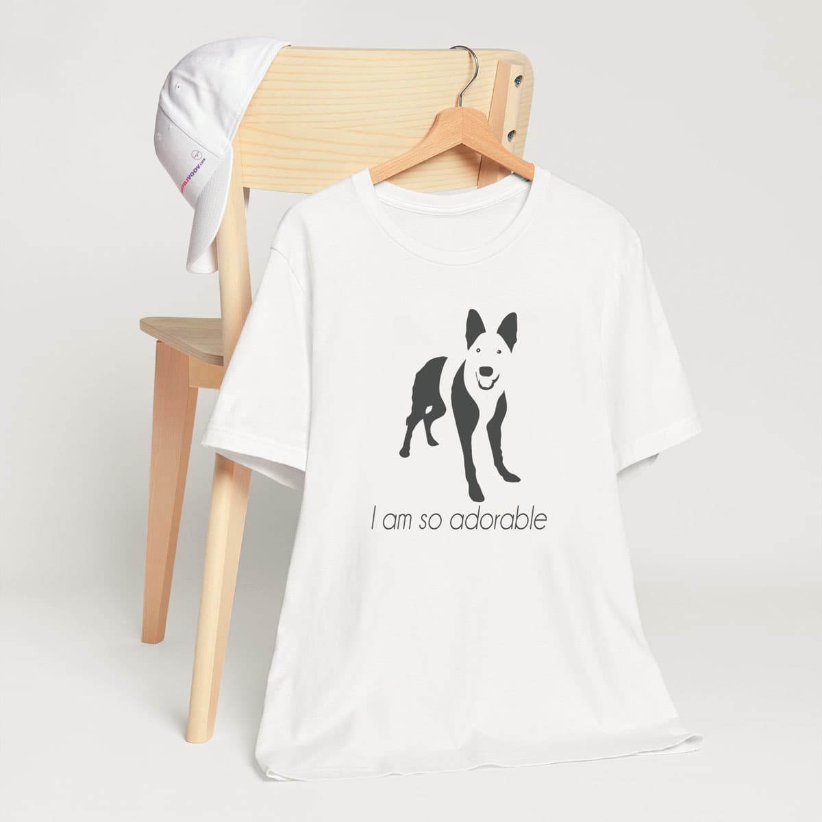 
White crew neck short sleeved t-shirt, featuring a one-line graphic print of a hand-drawn German Shepherd with its tongue out, and the playful text "I am so adorable”T-shirt hanging on chair shown in close-up to highlight print and size.