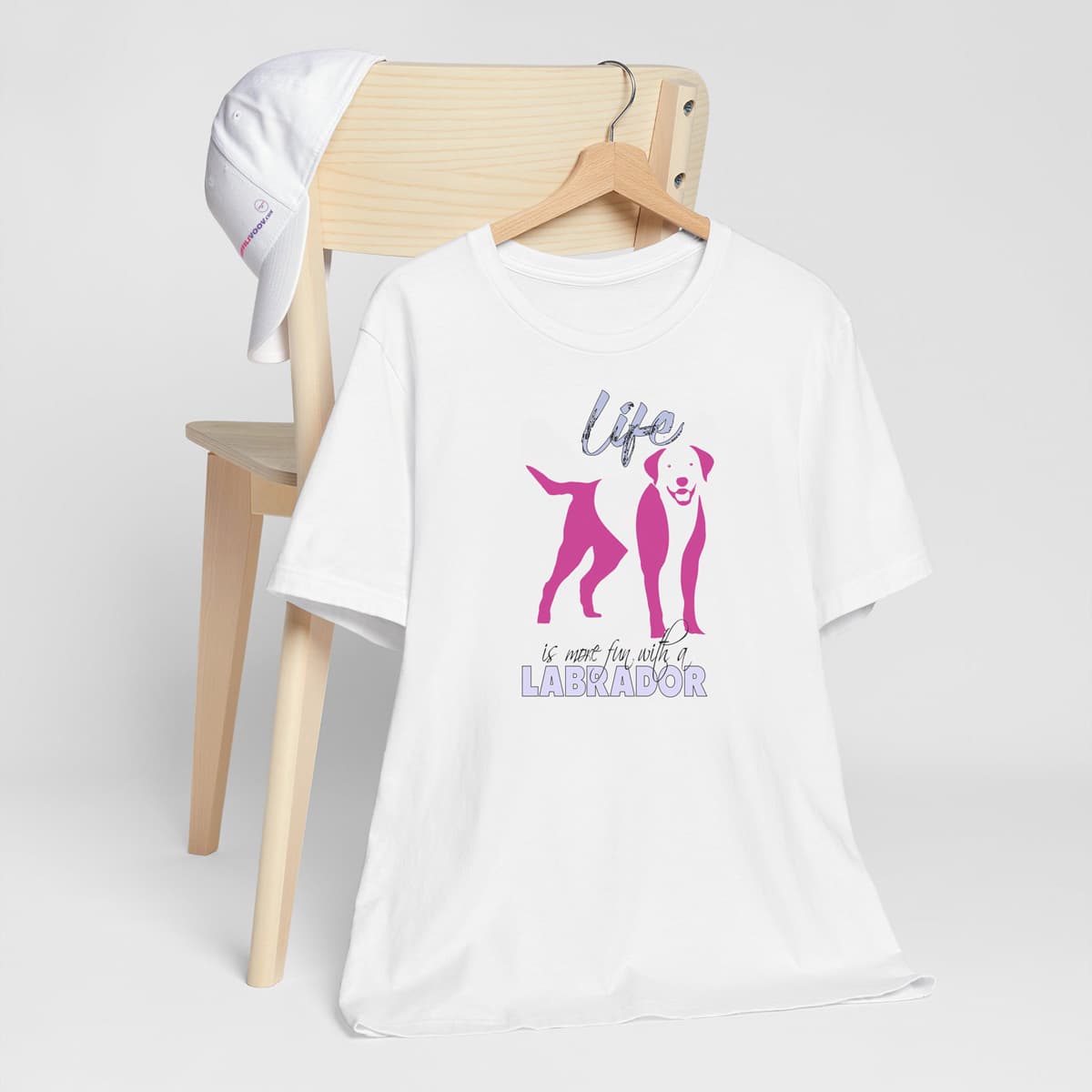 White crew neck short sleeved t-shirt. Featuring an adorable Labrador silhouette in cute pink, the tee also boasts a bold "Life" message and empowering "is more fun with a Labrador" phrase. T-shirt hanging on chair shown in close-up to highlight print and size