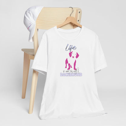 White crew neck short sleeved t-shirt. Featuring a silhouette Dachshund in vibrant pink, and quote with humor "life is more fun with a Dachshund"T-shirt hanging on chair shown in close-up to highlight print and size.