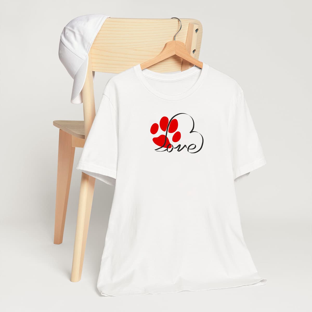 White crew neck short sleeved t-shirt, feature a heart-shaped graphic that transforms into the word "love" and a cute red paw. T-shirt hanging on chair shown in close-up to highlight print and size