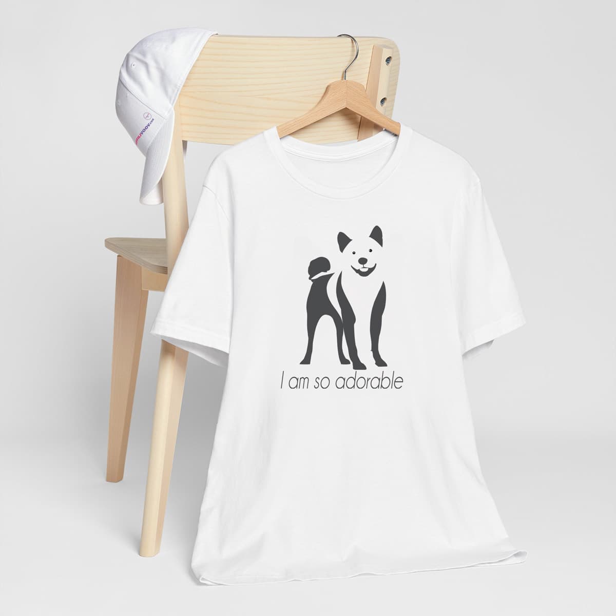 White crew neck short sleeved t-shirt, featuring a hand-drawn black and white one-line graphic print silhouette of an adorable Shiba Inu with its tongue out. The text "I am so adorable" underneath the dog .T-shirt hanging on chair shown in close-up to highlight print and size