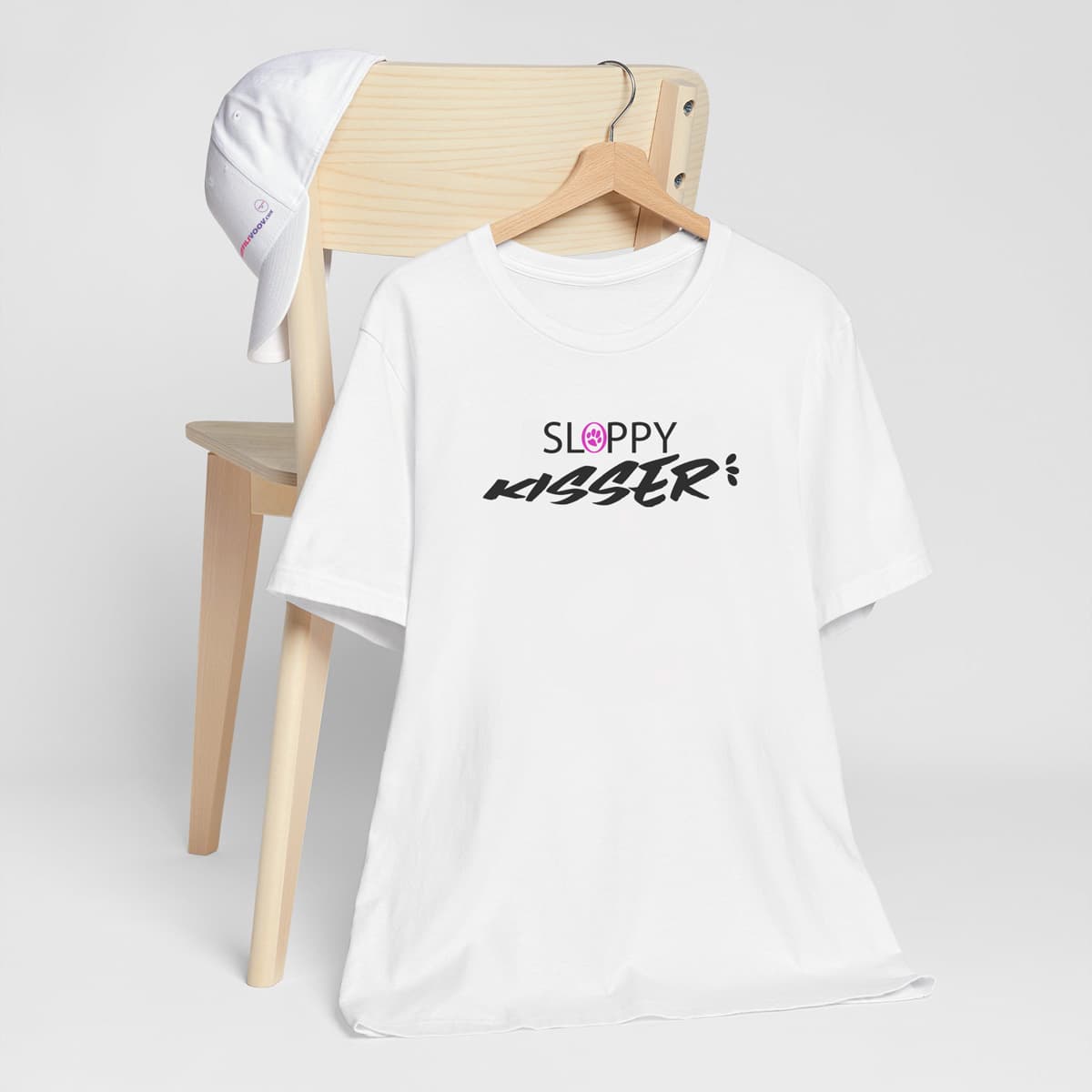 White crew neck short sleeved t-shirt, feature a funny message "sloppy kisser"with a pink logo paw, with a drool drops at the end of ”Sloppy kisser” text.  T-shirt hanging on chair shown in close-up to highlight print and size
