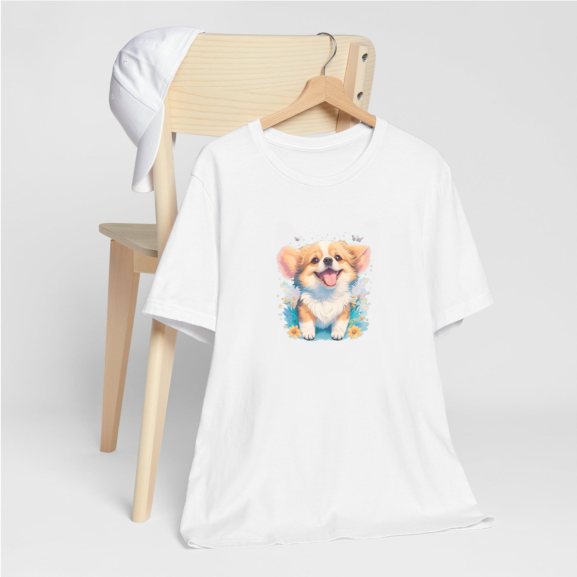 White crew neck short sleeved t-shirt,feature a graphic vibrant colorful hand-drawn dog face of a Corgi puppy sitting in floral garden.  T-shirt hanging on chair shown in close-up to highlight print and size