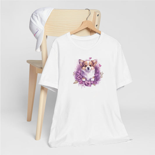 White crew neck short sleeved t-shirt, feature a graphic vibrant colorful hand-drawn Corgi puppy nestled amidst a garden of hearts and flowers in a charming wicker basket. T-shirt hanging on chair shown in close-up to highlight print and size.
