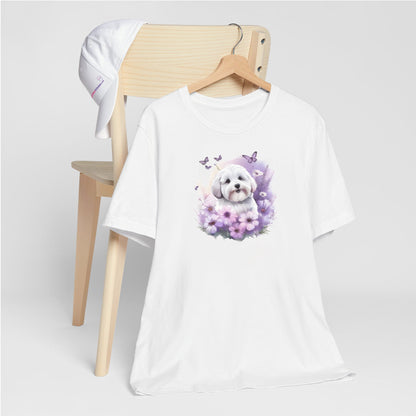 White crew neck short sleeve t-shirt with a colorful hand-drawn Coton de tulear puppy graphic sitting in a floral garden. shirt hanging on chair, close up showing bold dog art, cute pet-themed fashion