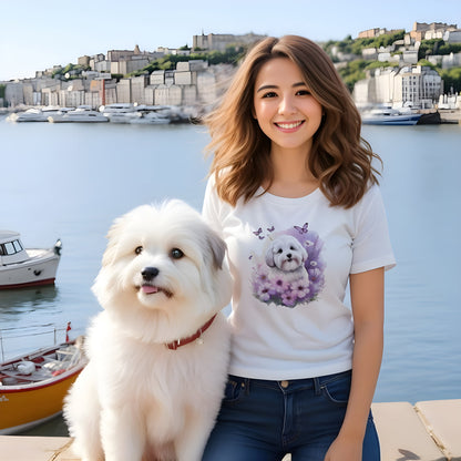 Women sitting at a harbor, close to her a coton de tulear puppy, she is wearing a white crew neck short sleeve t-shirt with a colorful hand-drawn Coton de tulear puppy graphic sitting in a floral garden. 