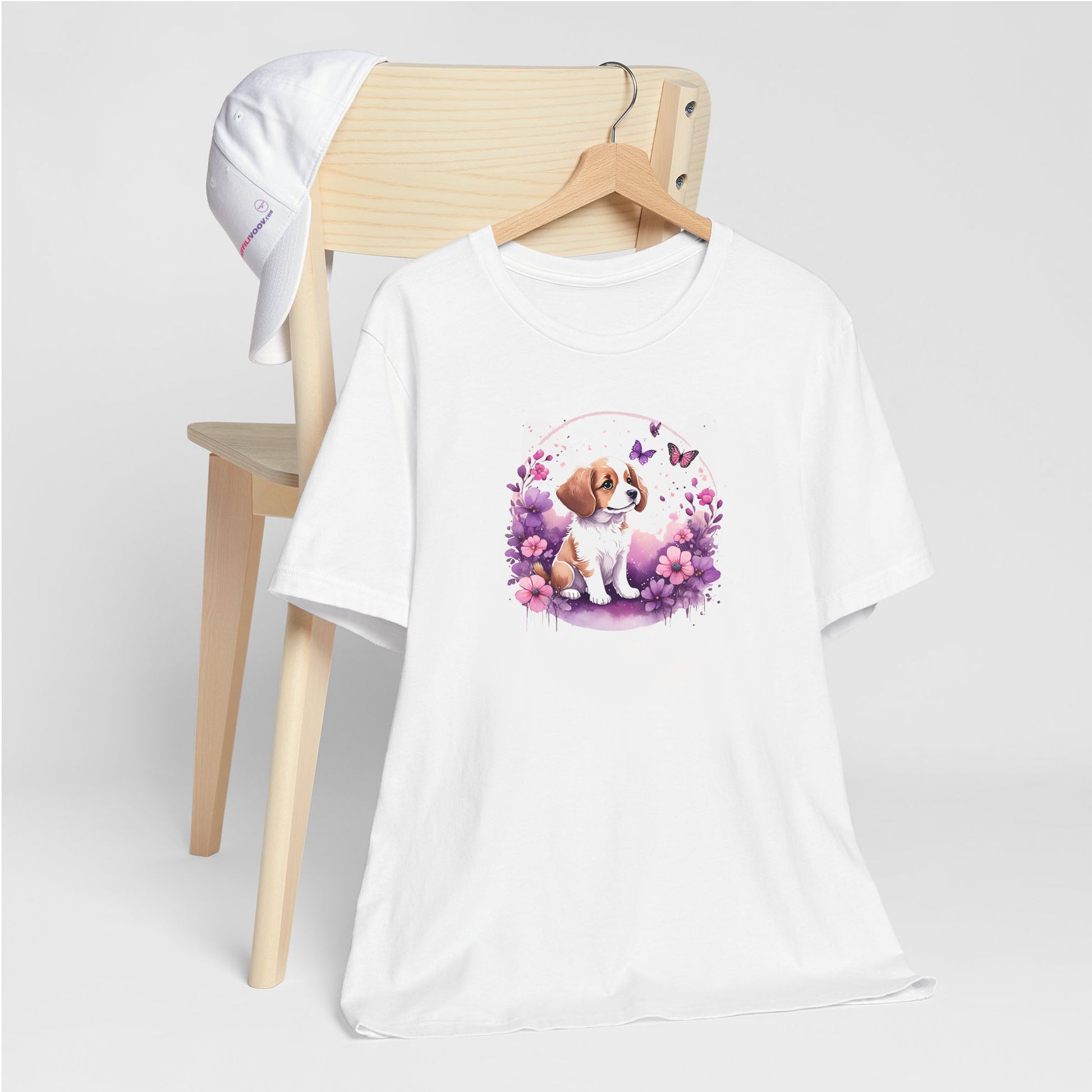 White crew neck short sleeved t-shirt, feature a playful pup amidst a whimsical floral fiesta. T-shirt hanging on chair shown in close-up to highlight print and size