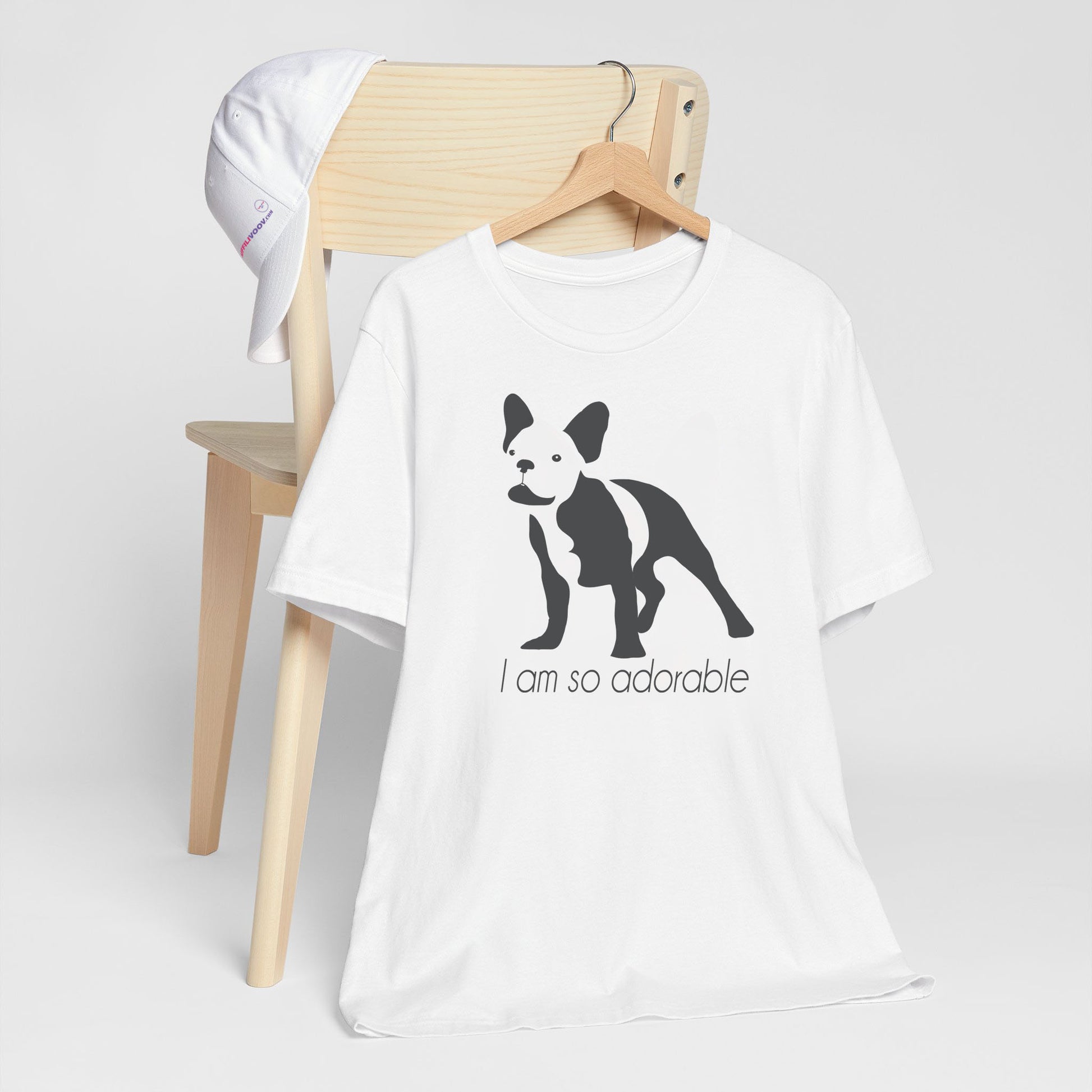 White crew neck short sleeved t-shirt, featuring a one-line graphic print of a hand-drawn French Bulldog with its tongue out, and the playful text "I am so adorable”. T-shirt hanging on chair shown in close-up to highlight print and size.