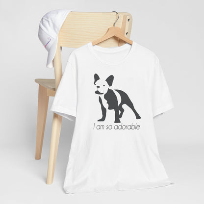 White crew neck short sleeved t-shirt, featuring a one-line graphic print of a hand-drawn French Bulldog with its tongue out, and the playful text "I am so adorable”. T-shirt hanging on chair shown in close-up to highlight print and size.